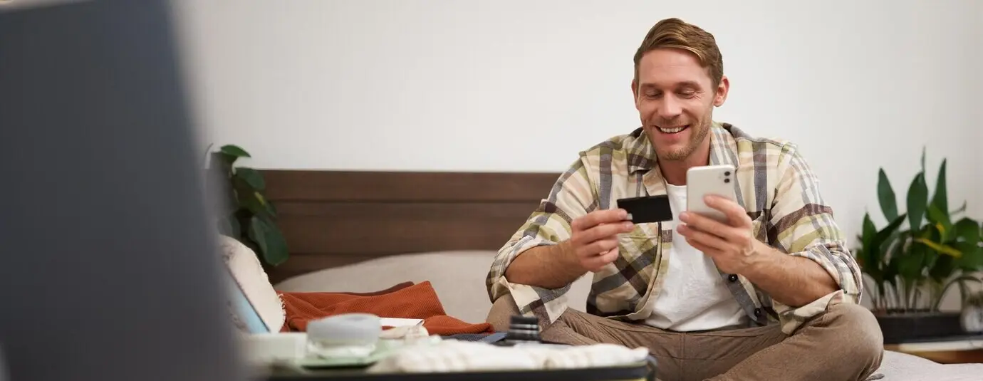 Портрет молодого мужчины, бронирующего билеты в отпуск, держащего кредитную карту и смартфон, сидящего с мобильным телефоном рядом с чемоданом, заполненным одеждой и готовым к отпускной поездке.