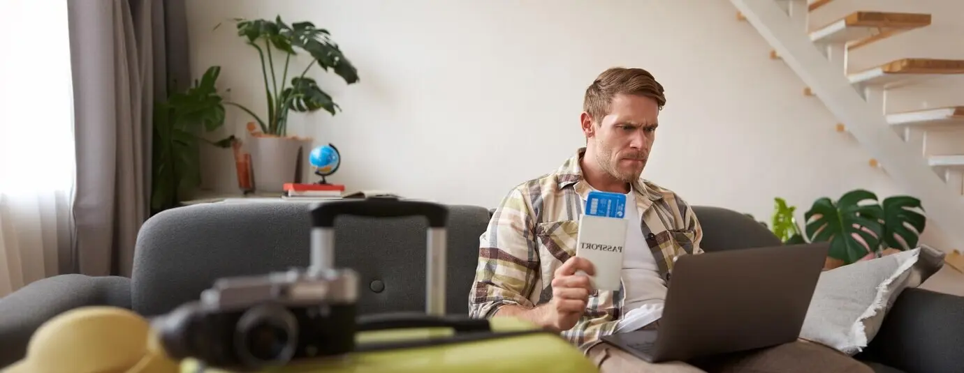 Сердитый мужчина с авиабилетами в руках недовольно смотрит на экран ноутбука — у него проблемы с его отпускным туром.